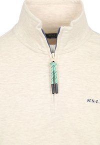 Lichtbeige fleece trui met een staande kraag, voorzien van een halve rits aan de voorkant en contrasterende groene en zwarte ritsen. Subtiele textuur en logo-details.