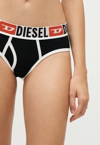 Μαύρα και άσπρα εσώρουχα Diesel που φοριούνται από ένα άτομο που δείχνει το κάτω μέρος του κορμού και το πάνω μέρος των μηρών σε ουδέτερο φόντο.