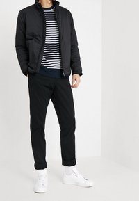 Giacca a vento nera con colletto alto, indossata sopra una camicia a righe blu navy e bianche, abbinata a pantaloni neri a sigaretta e sneakers bianche.