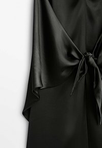 Tissu en satin noir avec un détail de nœud noué créant des plis drapés sur un fond clair uni.