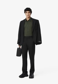Giacca nera sopra una camicia verde scuro con bottoni, abbinata a pantaloni neri e sneakers. Tiene in mano una piccola borsa nera. Sfondo bianco uniforme.
