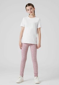 T-shirt bianco in cotone con maniche corte abbinato a leggings rosa con motivi floreali bianchi. Il modello indossa sneakers bianche.