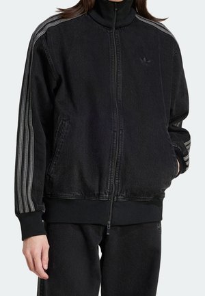 Persoon draagt een zwarte sportjas met rits en grijze strepen op de mouwen en bijpassende zwarte broek, hand gedeeltelijk in de zak.