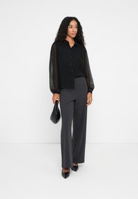 Blouse noire avec des manches bouffantes transparentes texturées, bouton sur le devant. Associée à un pantalon large taille haute gris et des talons noirs, tenant un sac à main.