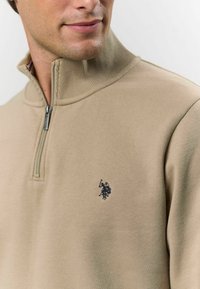 Béžová mikina na zips s vysokým golierom, ktorá má malé čierne vyšívané logo na hrudi a príjemnú, hladkú textúru.