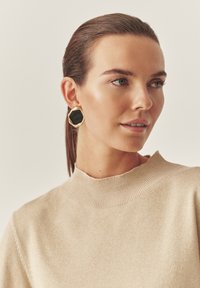 Czarno-złote ośmiokątne kolczyki noszone przez kobietę, połączone z jasnobeżowym swetrem z wysokim kołnierzem z prążkowanymi detalami.