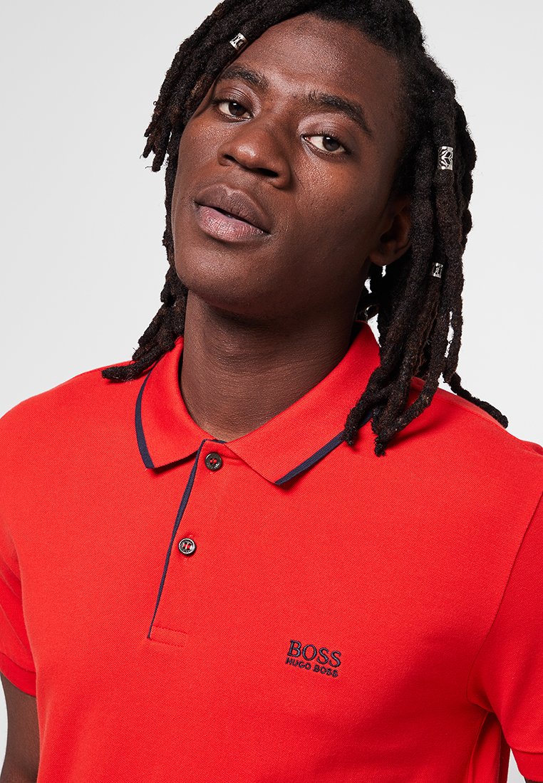 Polo rouge avec des accents bleu marine, manches courtes et un plastron à trois boutons. Arborant un logo sur la partie gauche de la poitrine.