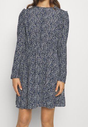 Vrouw die een marineblauwe jurk met lange mouwen draagt met een wit bloemenpatroon en een geplooide taille, staand tegen een effen achtergrond.