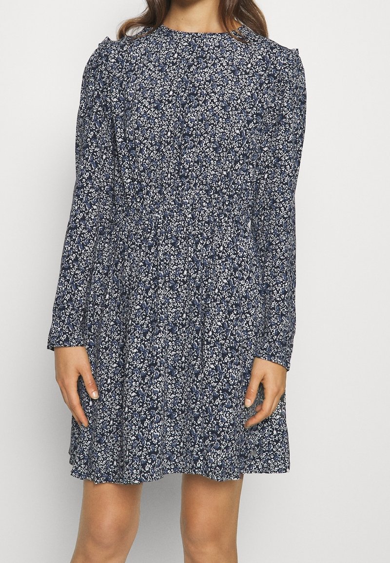 Femme portant une robe bleu marine à manches longues avec un motif floral blanc et une taille froncée, se tenant devant un fond uni.