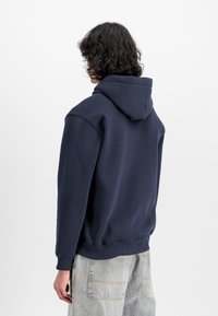 Felpa blu navy in cotone misto con tasca a marsupio, polsini a coste e cappuccio regolabile. Indossata con jeans grigi chiari.