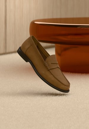 Mocassin en daim marron avec semelle noire flottant au-dessus d'un tapis beige clair, à côté d'un pied de table rond marron et brillant.