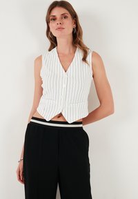 Bela pruga prsluka bez rukava z v-izrezom, pet gumbov spredaj in prilagojen kroj. Kombiniran z črnimi hlačami z prugastim pasom.