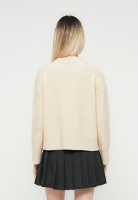 Personne avec de longs cheveux ombrés portant un pull beige et une jupe plissée gris foncé, de dos devant un fond uni.