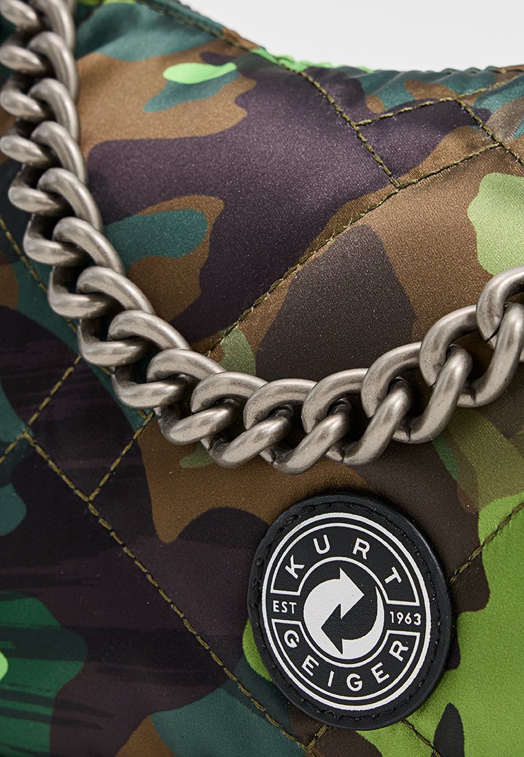 Gequiltte tas met een camouflagepatroon in groen, bruin en zwart. Zilveren schakelsierraad en een ronde logopatch. Gemosseerde afwerking.