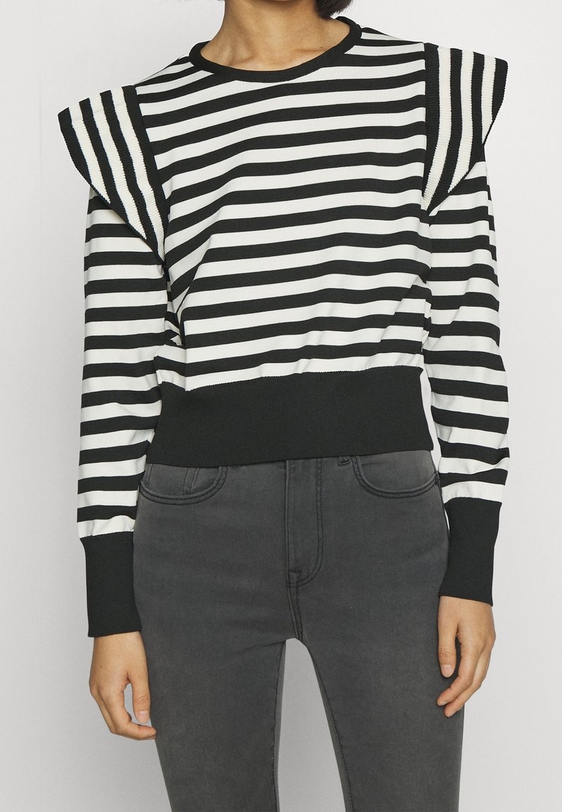 Il maglione a righe bianche e nere presenta dettagli arricciati sulle spalle, scollatura rotonda e polsini a costine con una vita aderente. Abbinato a jeans grigi.
