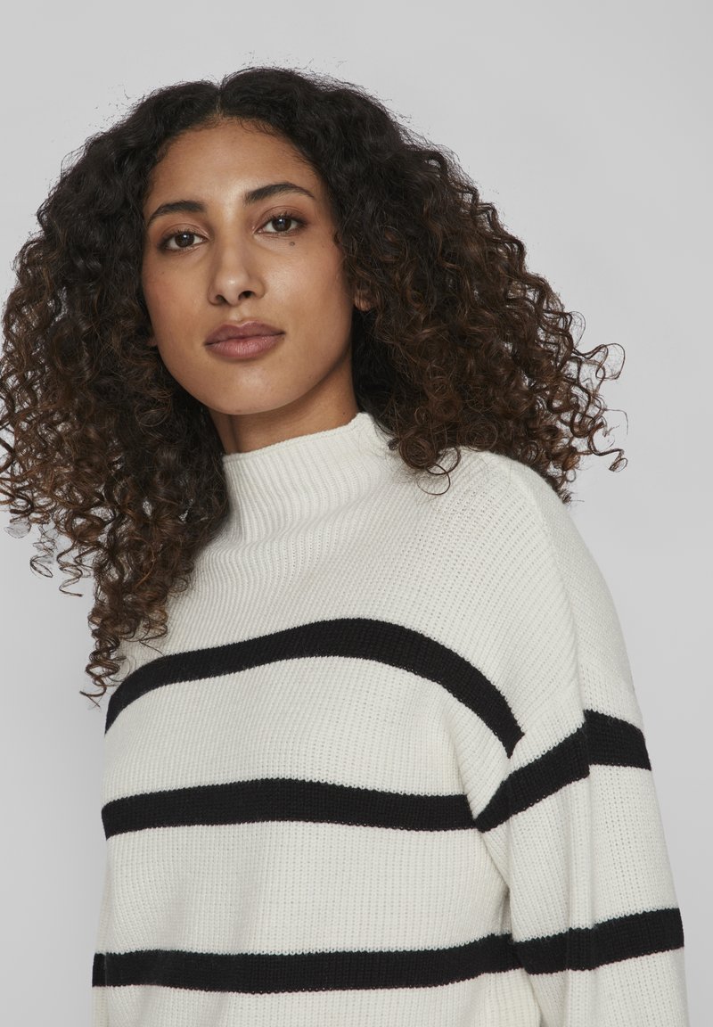 Pull en tricot crème avec des rayures horizontales noires, un col montant et une finition texturée. Le design est décontracté et surdimensionné.