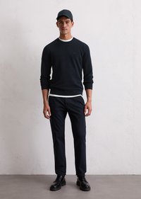 Pulover tricotat bleumarin cu detalii albe la guler, purtat cu pantaloni închiși la culoare și cizme negre cu șireturi, împreună cu o șapcă asortată. Stând în fața unui zid deschis.