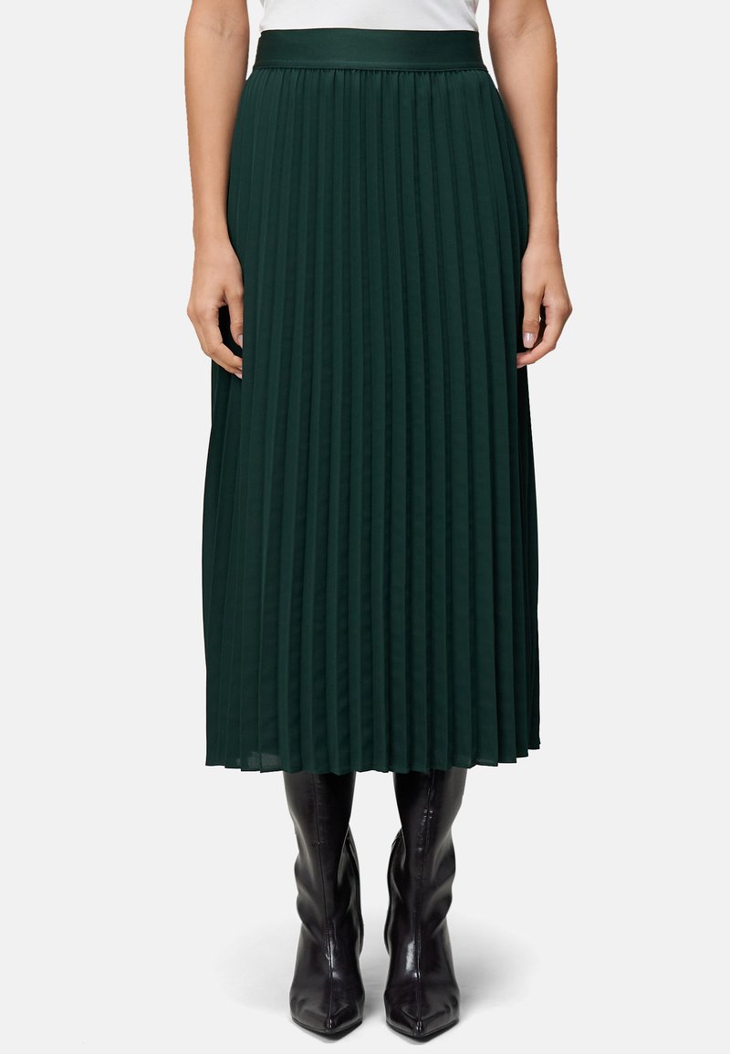 Jupe midi plissée verte avec une taille large, en tissu léger, associée à des bottes noires à bout pointu. Texture verticale visible.
