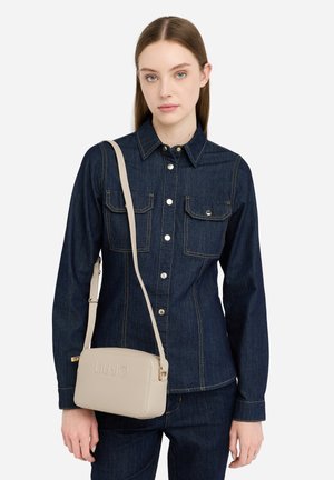 Beige crossbody bag with embossed logo, smooth texture, rectangular shape, and gold hardware. Worn with a dark denim shirt.