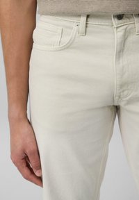 Pantalones vaqueros de mezclilla beige claro con un diseño ajustado, estilo de cinco bolsillos y una tela texturizada sutil. Presenta un botón de metal y un acolchado mínimo.