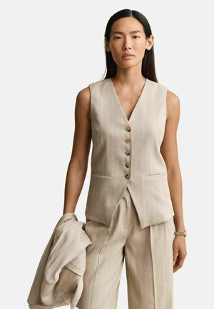 Frau trägt eine beige Nadelstreifenweste und passende weit geschnittene Hosen, hält in ihrer linken Hand eine passende Jacke.