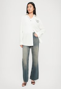 Žena stojí v bielej oversized košeli, vyblednutých úzkych džínoch a hnedých sandáloch na otvorenej špičke na neutrálnom svetlom pozadí.