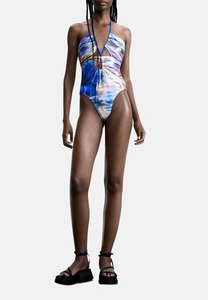 Naine kannab värvikat mustriga üheosalist ujumistrikood lõigetega, mustade platvormsandaalidega, seisab lihtsa tausta ees.