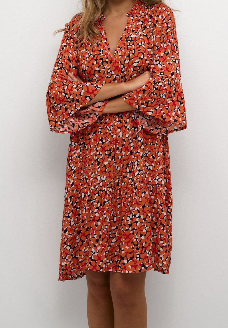 Femme portant une robe orange longueur genoux avec un motif floral rouge, noir et blanc et des manches larges évasées, debout, les bras croisés.