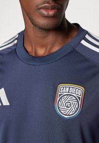 Camiseta atlética en color navy con cuello acanalado, que presenta rayas blancas en los hombros y un logo bordado de "San Diego FC" en el pecho.