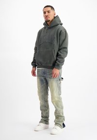 Donkergroene hoodie met voorzak en reliëf logo, gecombineerd met lichtblauwe distressed jeans en witte sneakers. Model staat tegen een effen achtergrond.