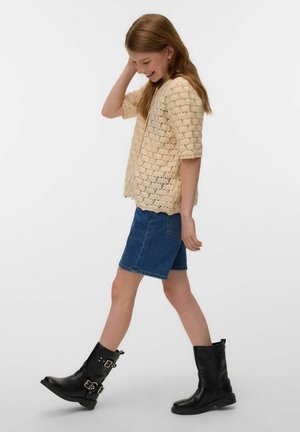 Niña con cabello largo caminando, vestida con un top corto de crochet beige, falda azul de mezclilla y botas negras con hebilla sobre un fondo blanco.