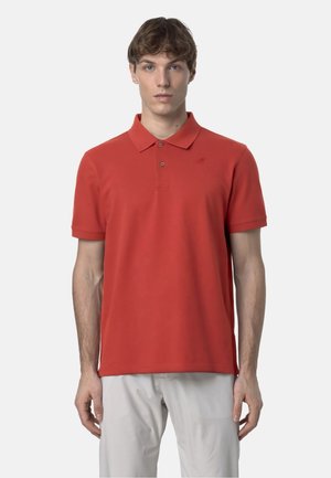 Uomo con capelli castani corti che indossa una polo rossa a maniche corte e pantaloni grigio chiaro, in piedi davanti a uno sfondo chiaro e semplice.