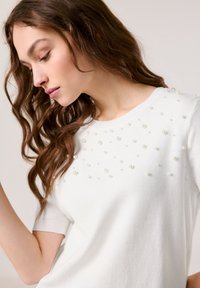 Weißer Strickpullover mit Rundhalsausschnitt und kurzen Ärmeln, verziert mit verstreuten Kunstperlen an der Vorderseite für zusätzliche Textur.