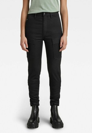 Kobieta ubrana w czarne spodnie cargo z wysokim stanem i kieszeniami z klapami oraz czarne botki na masywnej podeszwie, stojąca na białym tle.