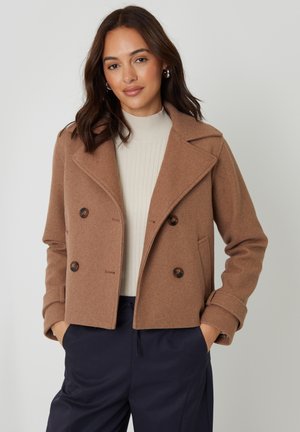 Kobieta o brązowych włosach do ramion, ubrana w dwurzędowy płaszcz koloru wielbłądziego z białym, prążkowanym golfem, ręce w kieszeniach granatowych spodni.