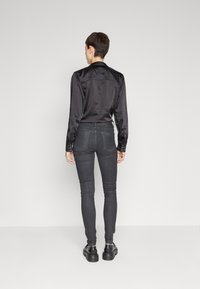 Camisa de satén negra con un diseño ajustado y puños abotonados, combinada con jeans oscuros ajustados y zapatos negros. Vista trasera mostrada.