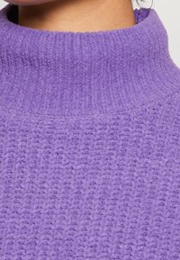 Neulottu pooloneule, valmistettu teksturoidusta purppurasta kankaasta. Siinä on resorinen pääntie ja pehmeä, kodikas ulkonäkö.