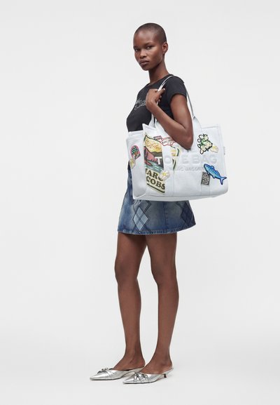 Femme en t-shirt noir, jupe en jean et ballerines argentées, debout tenant un grand sac cabas blanc orné de patchs colorés et de motifs graphiques.