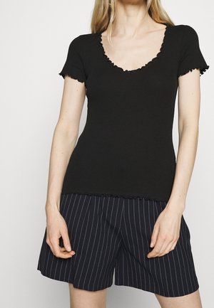 Femme portant un haut noir à manches courtes avec des bords froncés et un short noir à fines rayures, debout devant un fond uni.