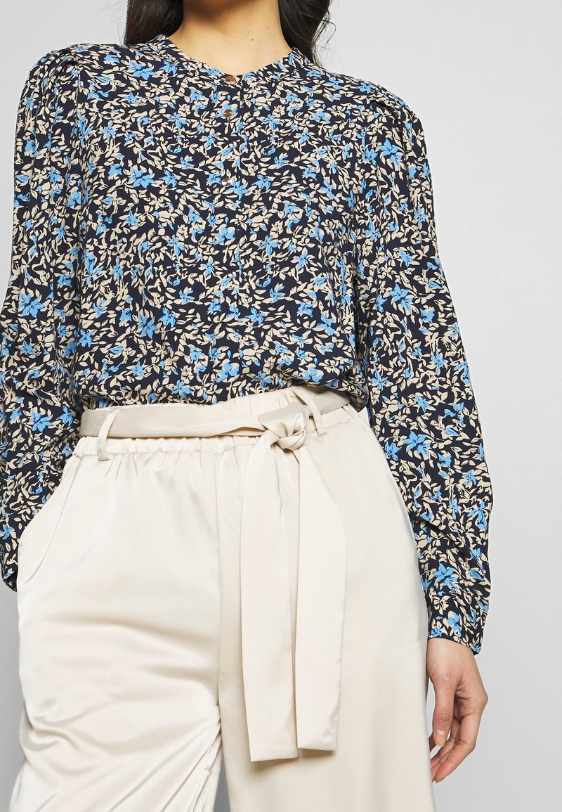 Kwiatowa bluzka w kolorze granatowym z niebieskimi i beżowymi wzorami, z okrągłym dekoltem i długimi rękawami, zestawiona z kremowymi spodniami z paskiem.