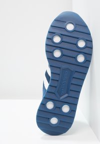 Modrá gumová podrážka tenisky s bílými akcenty. Obsahuje texturovaný grip, kulaté podložky a vroubkovaný design. Branding viditelný na podrážce.