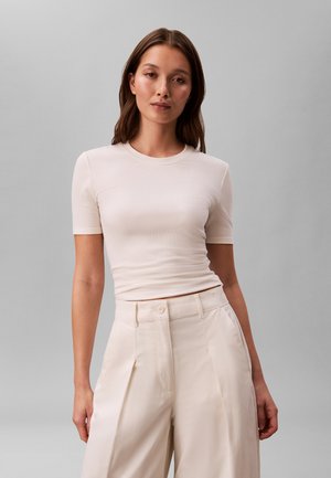 Femme aux longs cheveux bruns portant un haut beige ajusté à manches courtes et un pantalon beige large taille haute, sur un fond gris uni.