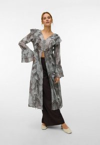 Femme portant une tunique transparente à manches longues à volants avec un motif, sur un pantalon noir large et des chaussures crème à petits talons, posant avec une main sur la hanche.