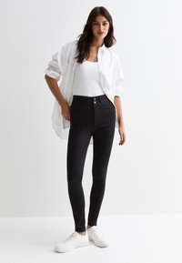 Jean noir taille haute avec une texture lisse, fermeture à bouton et poches avant, associé à un débardeur blanc et une chemise blanche surdimensionnée.