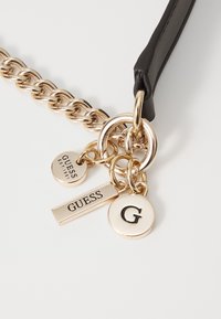Schwarzes Lederarmband mit goldfarbenem Metallchain, ausgestattet mit drei kreisförmigen und rechteckigen Anhängern, die mit "GUESS" und "G." beschriftet sind.