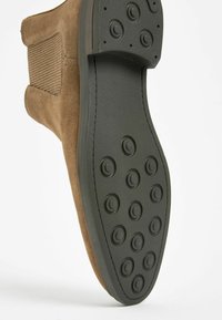 Bota de ante marrón con paneles laterales elásticos acanalados, puntera redondeada y suela de goma negra texturizada con patrones de agarre circulares.