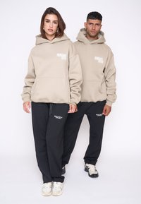 Sudaderas beige con bolsillo frontal y logotipo, combinadas con pantalones deportivos negros que presentan un logotipo. El calzado del modelo incluye zapatillas de colores claros.