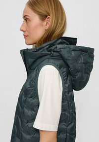 Puffige, dunkelgrüne ärmellose Weste mit Kapuze, die über einem weißen Kurzarmshirt getragen wird und einen gesteppten Stoff sowie einen Reißverschluss hat.