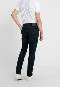 Dark denim trousers with a slim fit, featuring a black waistband and pockets. Paired with a plain white t-shirt and white sneakers.