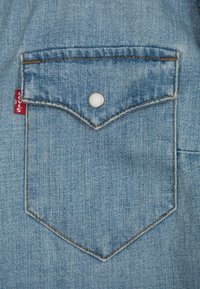 Poche en denim bleu clair avec un bouton-pression blanc, des bords surpiqués et une étiquette rouge sur le côté, avec un rabat pointu.